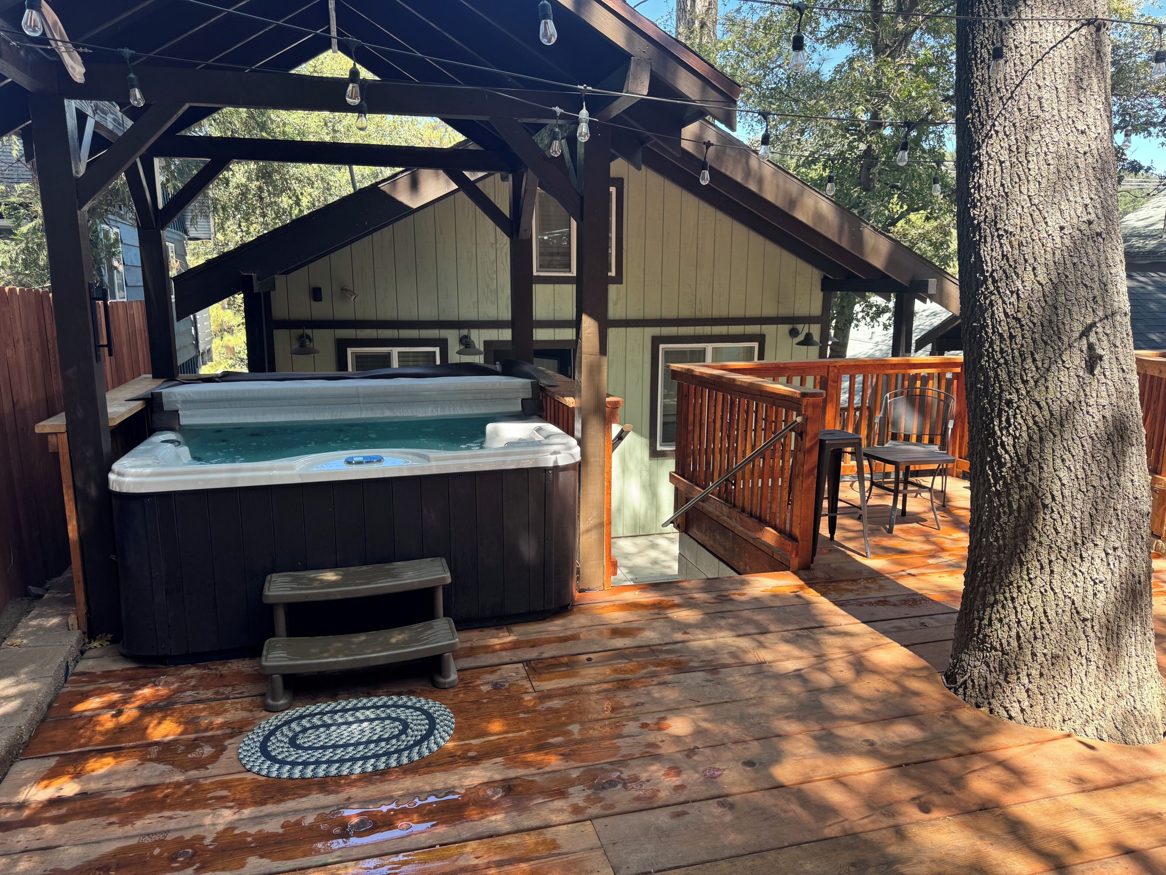A hot tub under a wooden pergola with a wooden deck, a wooden fence, and a tree.