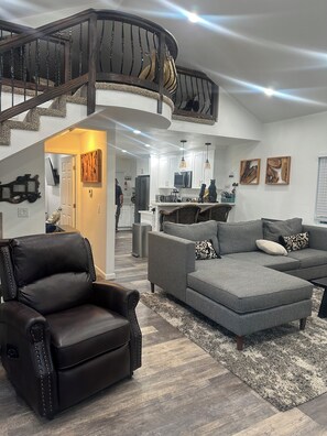 A modern living room with a leather armchair, grey sectional sofa, and a staircase with a wooden railing. - Charming 4-bedroom house walking distance to downtown Crestline and Lake Gregory (Crestline)