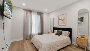A bedroom with a bed, a TV, a bedside table, a mirror, and a window with curtains.