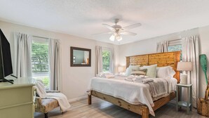 A bedroom with a wooden bed, a desk, a chair, a ceiling fan, and a window with curtains. - The Silver Shell SSI - Dog Friendly (St. Simons Island)
