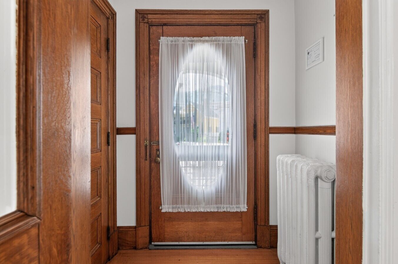 Una puerta de madera con una cortina blanca, un radiador y una pared blanca con adornos de madera.