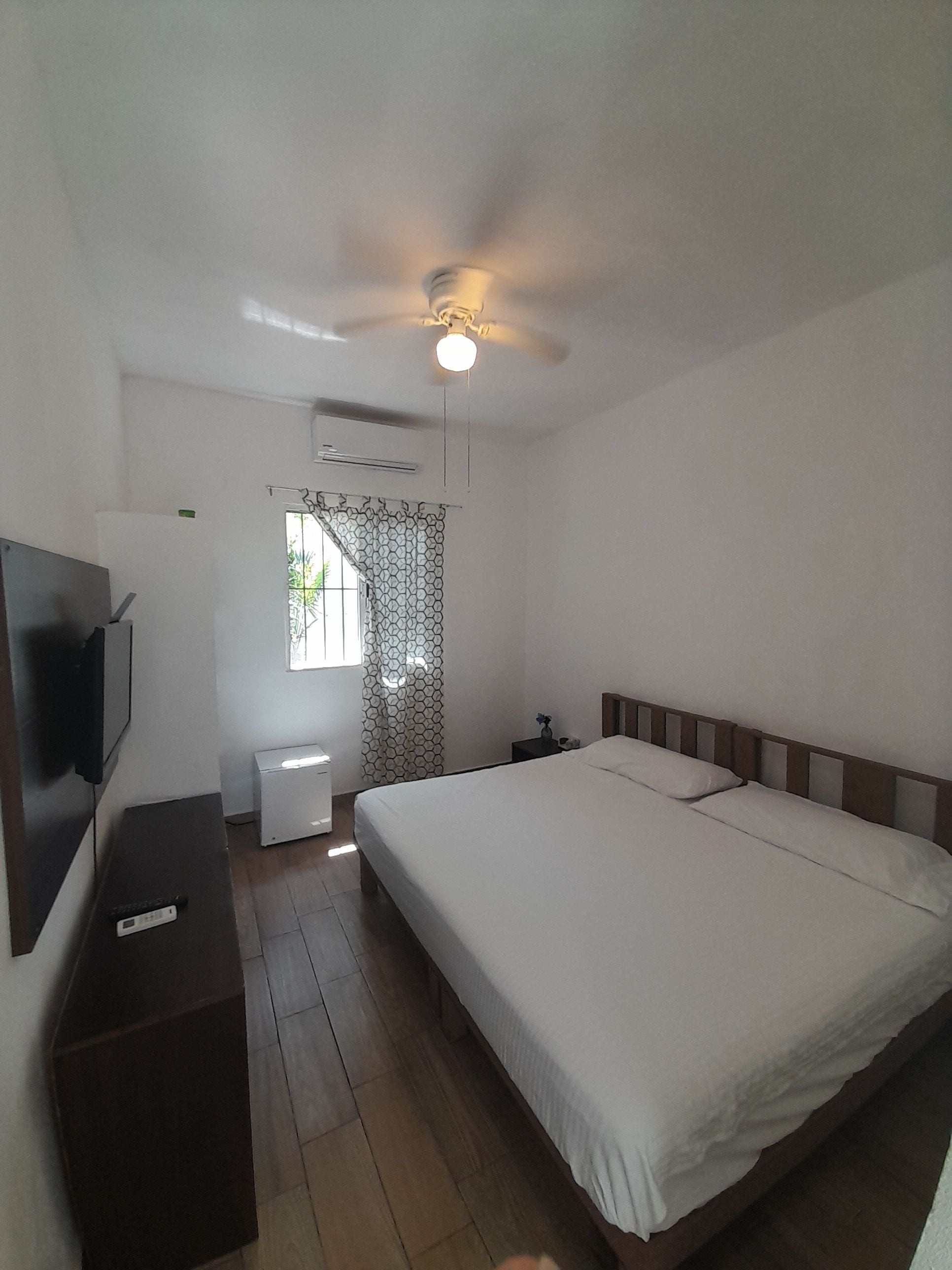A bedroom with a bed, a television, a ceiling fan, and a window with patterned curtains.