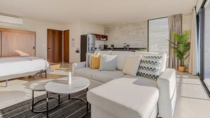 Una habitación de hotel moderna con cama, sofá, mesa de comedor, zona de cocina y ventanas grandes.