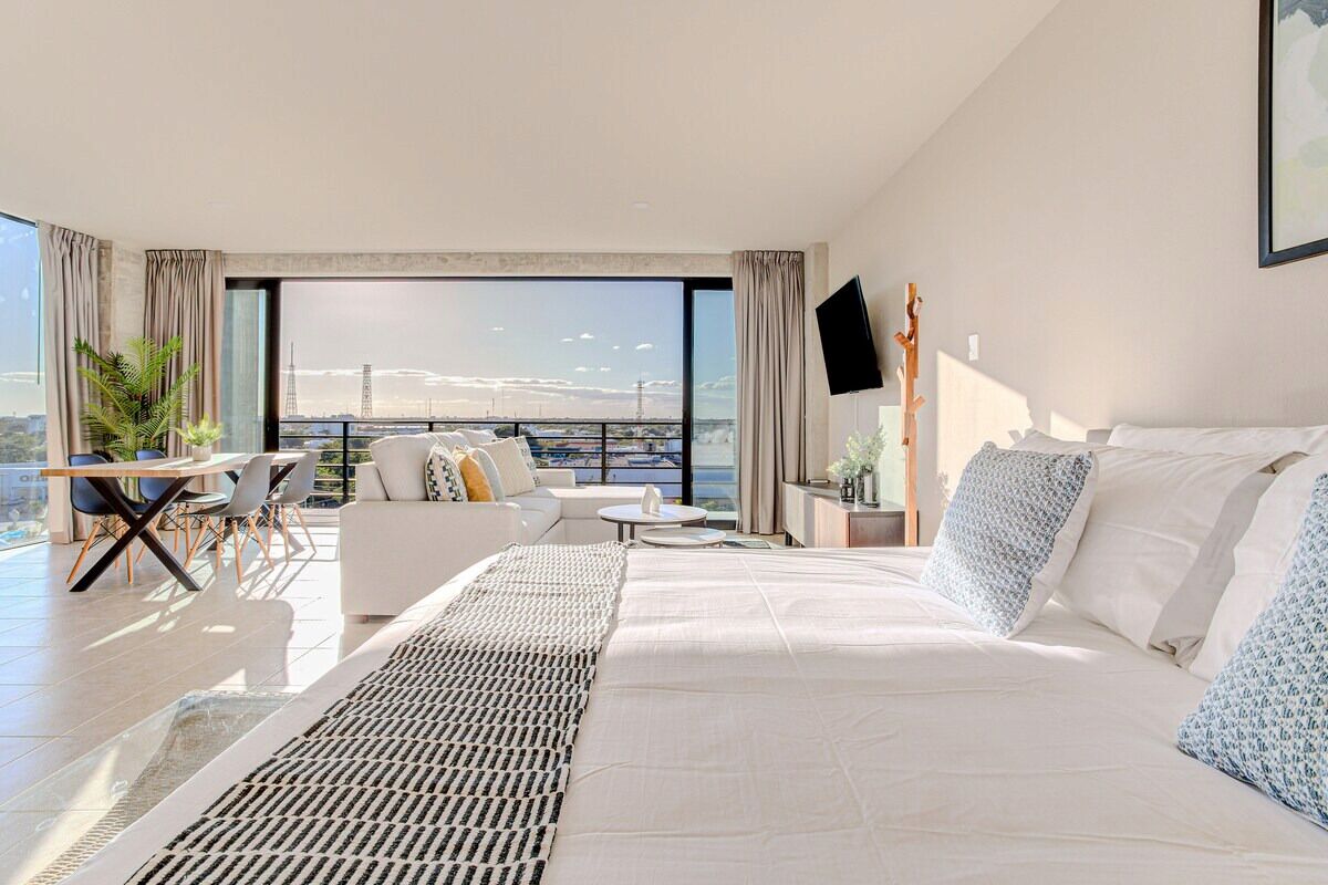 A modern hotel room with a large bed, a balcony with a view of the city, a desk, and a chair.