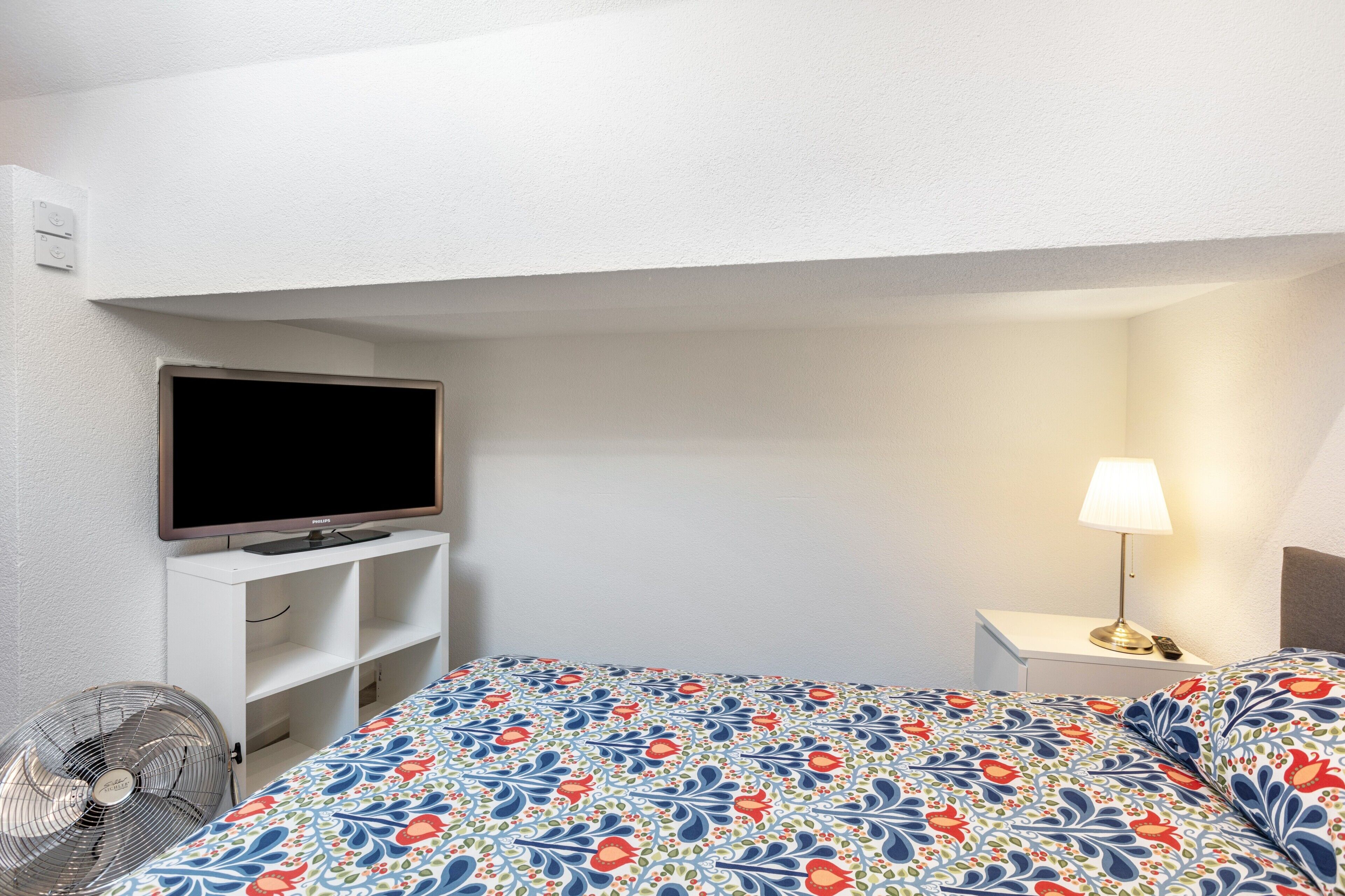 A hotel room with a bed, a TV, a bedside lamp, and a fan.