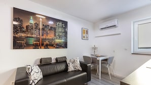 A modern living room with a black leather sofa, a dining table with two chairs, a cityscape painting, and a wall-mounted air conditioner.