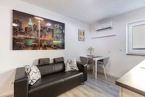 A modern living room with a black leather sofa, a dining table with two chairs, a cityscape painting, and a wall-mounted air conditioner.