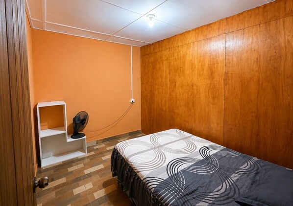 A bedroom with a bed, a fan, a shelf, and a wooden wall.