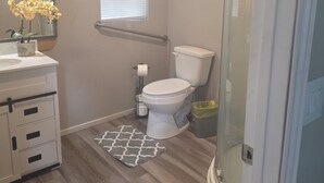 A bathroom with a toilet, sink, mirror, window, shower, and two rugs. - Cozy 1-bedroom cabin in serene Magnolia with AC (Magnolia)