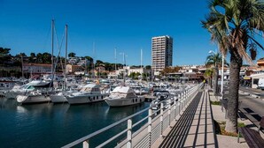 A marina with boats docked, a pedestrian walkway, and a tall building in the background. - In the heart of the Calanque du Cap Rousset—Apartment with a sea view, perfect for a romantic getaway! (Carry-le-Rouet)
