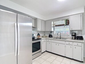 A modern kitchen with white cabinets, a stainless steel refrigerator, a built-in oven, a coffee maker, and a window with blinds. - The Jefferson 220 - Downtown Saint Pete (St. Petersburg)