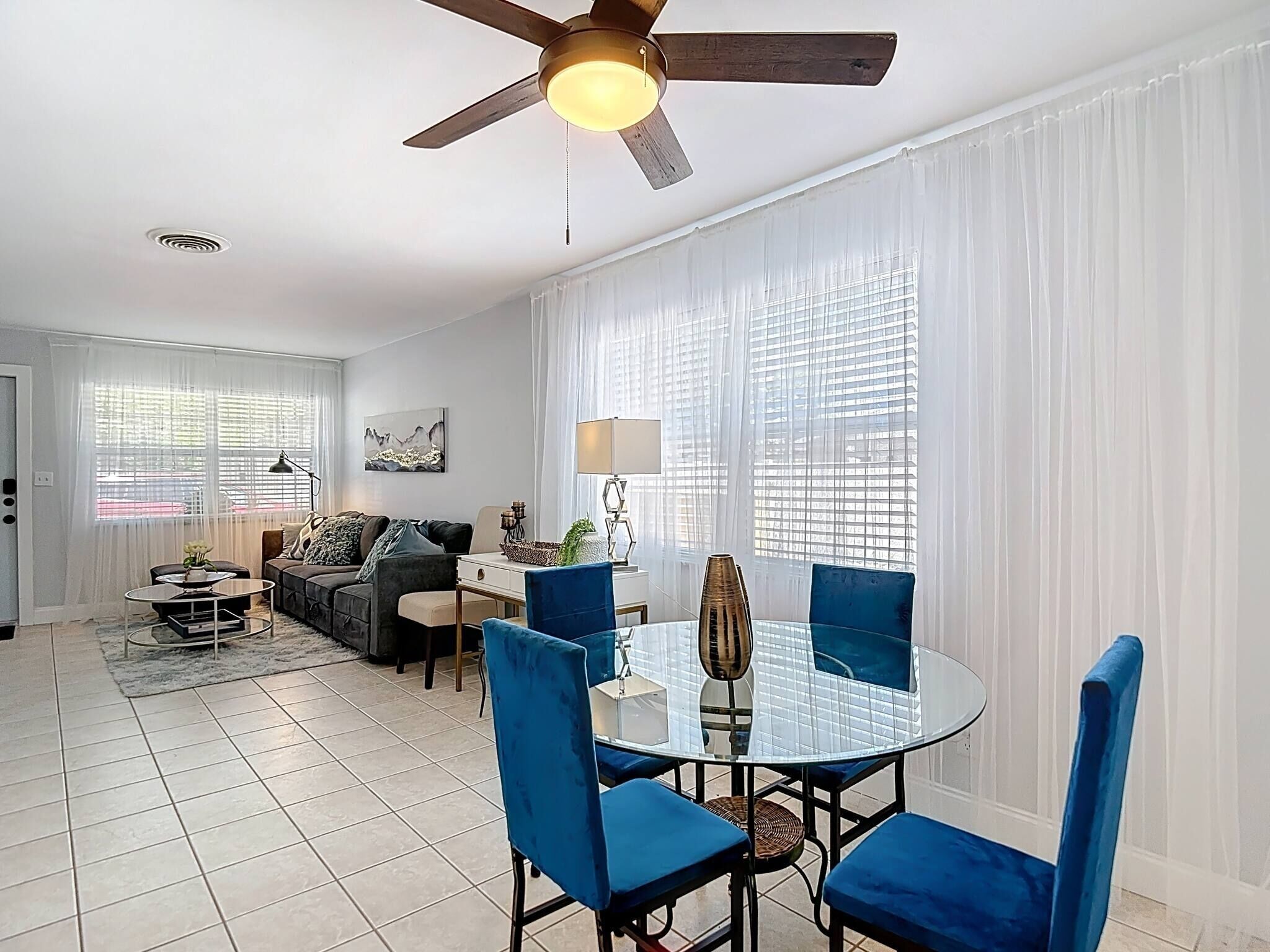 A living room with a ceiling fan, a dining area with a glass table and blue chairs, a sofa, and a lamp.