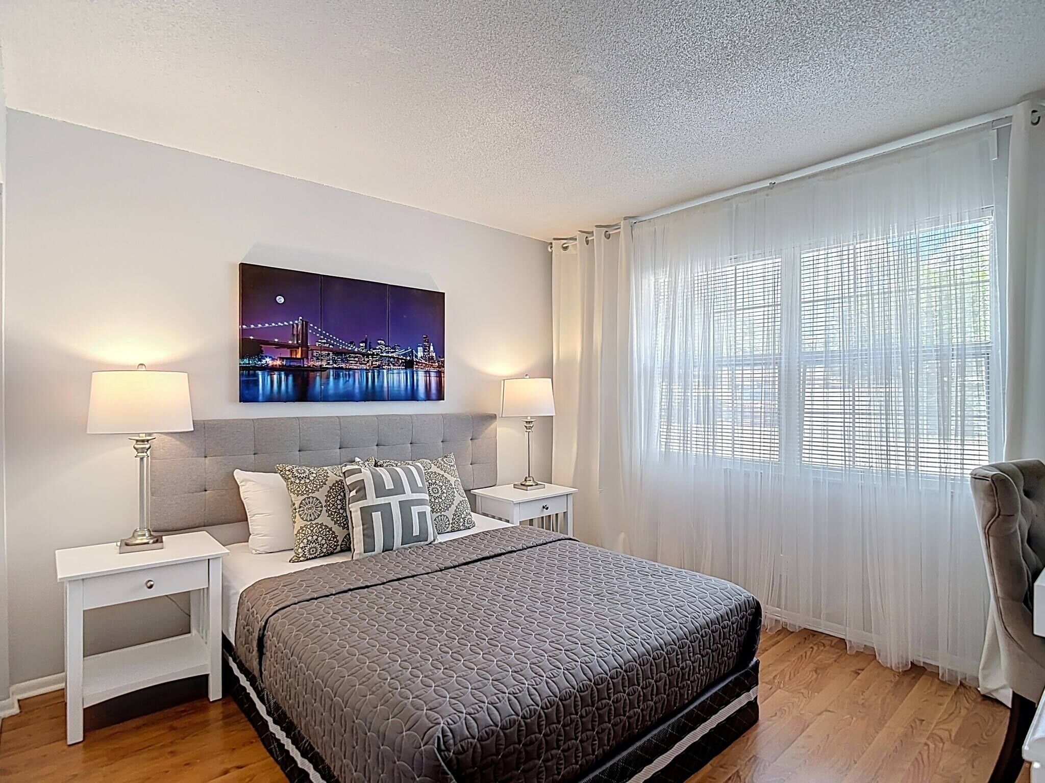 Un dormitorio con cama, dos mesitas de noche, una mesilla, una ventana con cortinas translúcidas y un cuadro de un paisaje urbano en la pared.