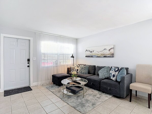 A living room with a sofa, a chair, a coffee table, a door, and a window with curtains. - The Jefferson 220 - Downtown Saint Pete (St. Petersburg)