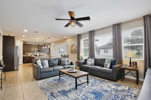 A modern living room with two grey sofas, a coffee table, a ceiling fan, and a kitchen in the background. - The Magic Haven - 5BR - Sleeps 13 - Near Disney (Davenport)