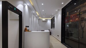 Zona de recepción de un hotel con un mostrador blanco, un punto de control de seguridad y una puerta de cristal hacia la entrada del hotel.