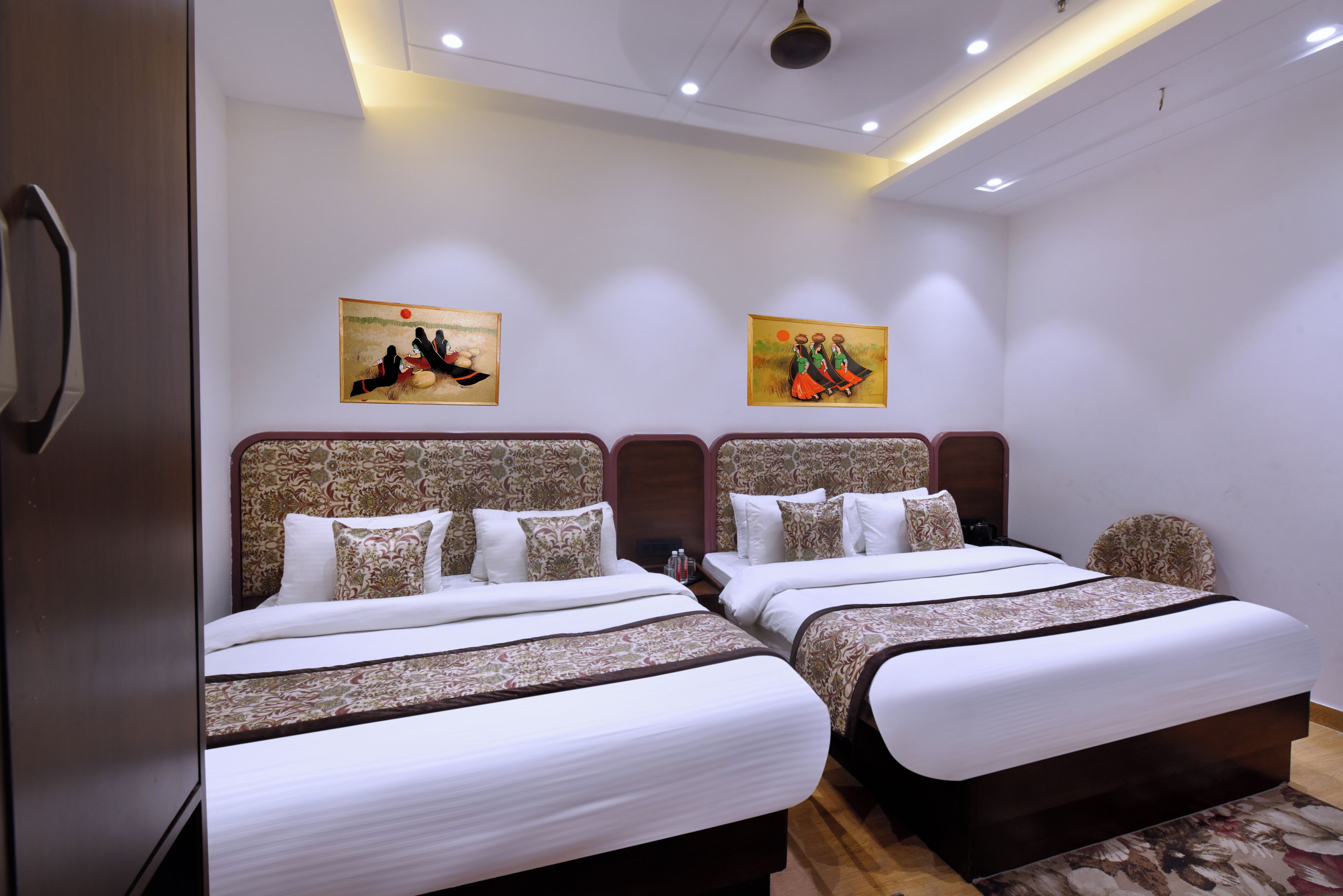 A hotel room with two beds, each with patterned headboards and white bedding. There are two wall-mounted paintings above the beds.