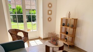 Uma sala de estar com uma cadeira de vime, um sofá, uma mesa redonda, uma estante de madeira e uma vista para um jardim através de uma porta de vidro.