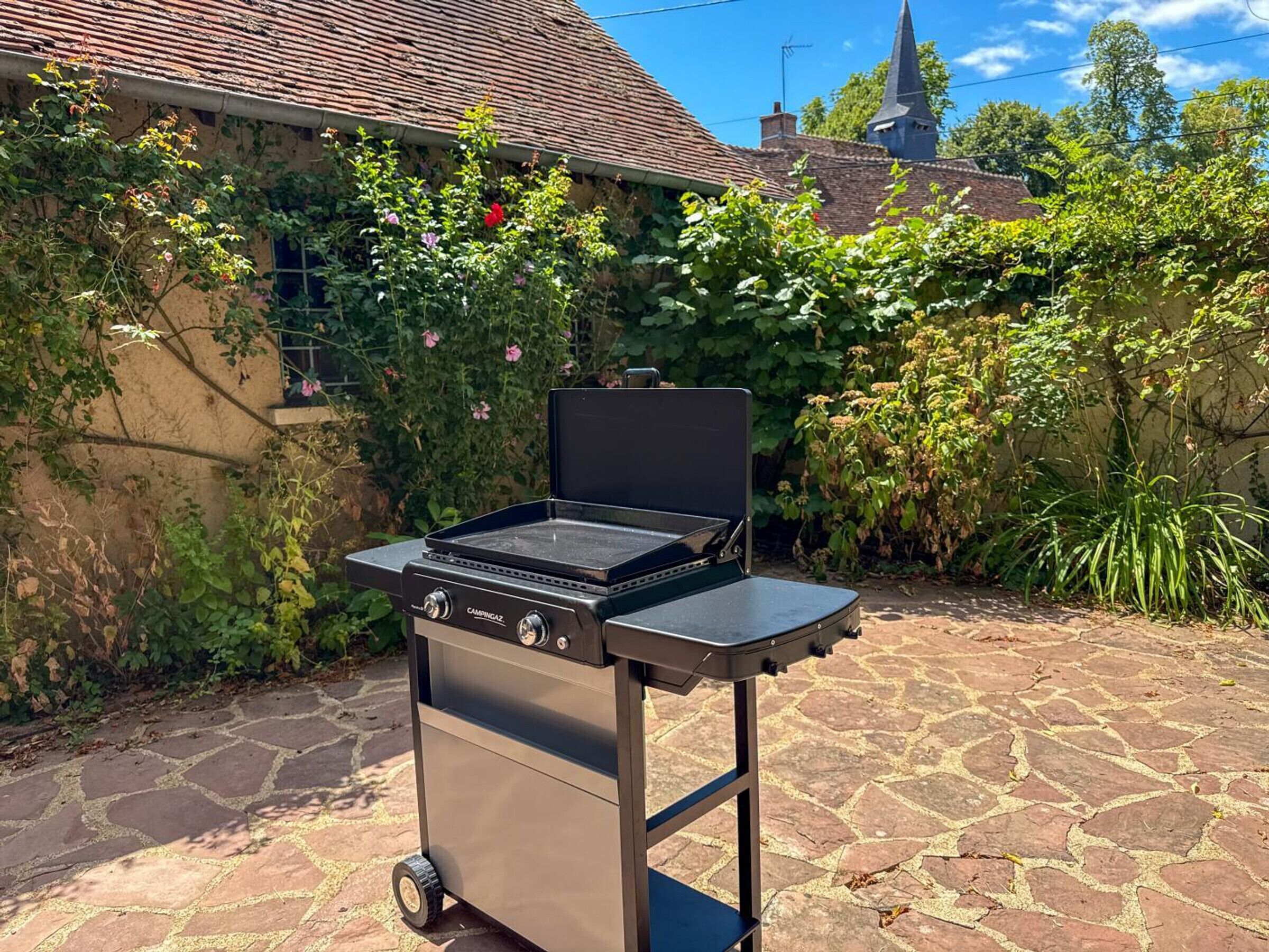 En sammenklappelig campinggrill på hjul, placeret på en stenbelagt terrasse.