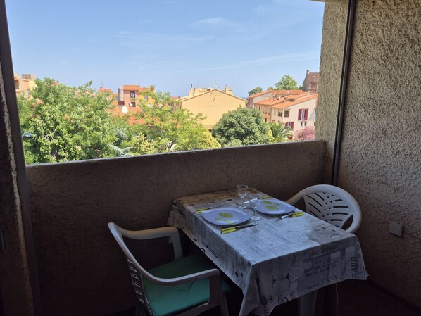 A balcony with a table set for two, a chair, and a view of buildings and trees. - Charming apartment, private parking - 2 guests (Collioure)