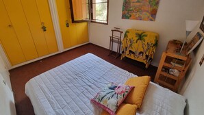 Ein Schlafzimmer mit einem Bett, einem Fenster, einem gelben Kleiderschrank, einem kleinen Tisch und einem Bild an der Wand.