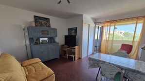 Ein Wohnzimmer mit einer Couch, einem Fernseher, einem Tisch mit Stühlen und Blick durch ein Fenster.