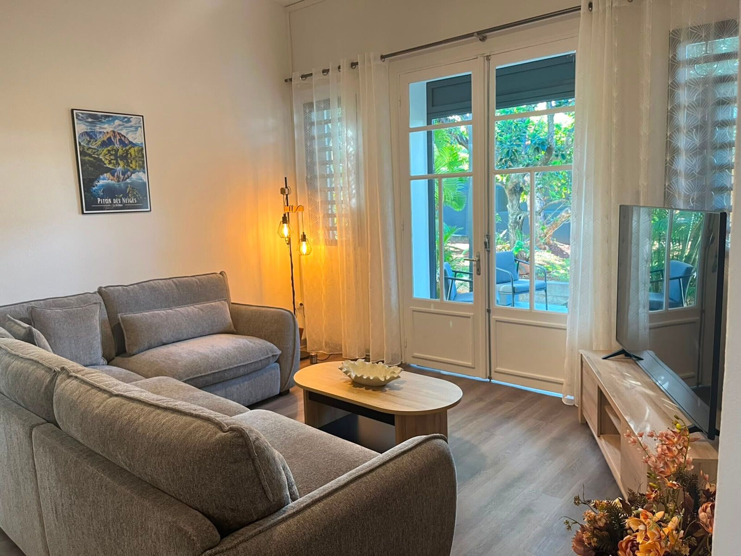 A living room with a sofa, a small table, a TV, and a large sliding glass door leading to an outdoor area with chairs and plants.