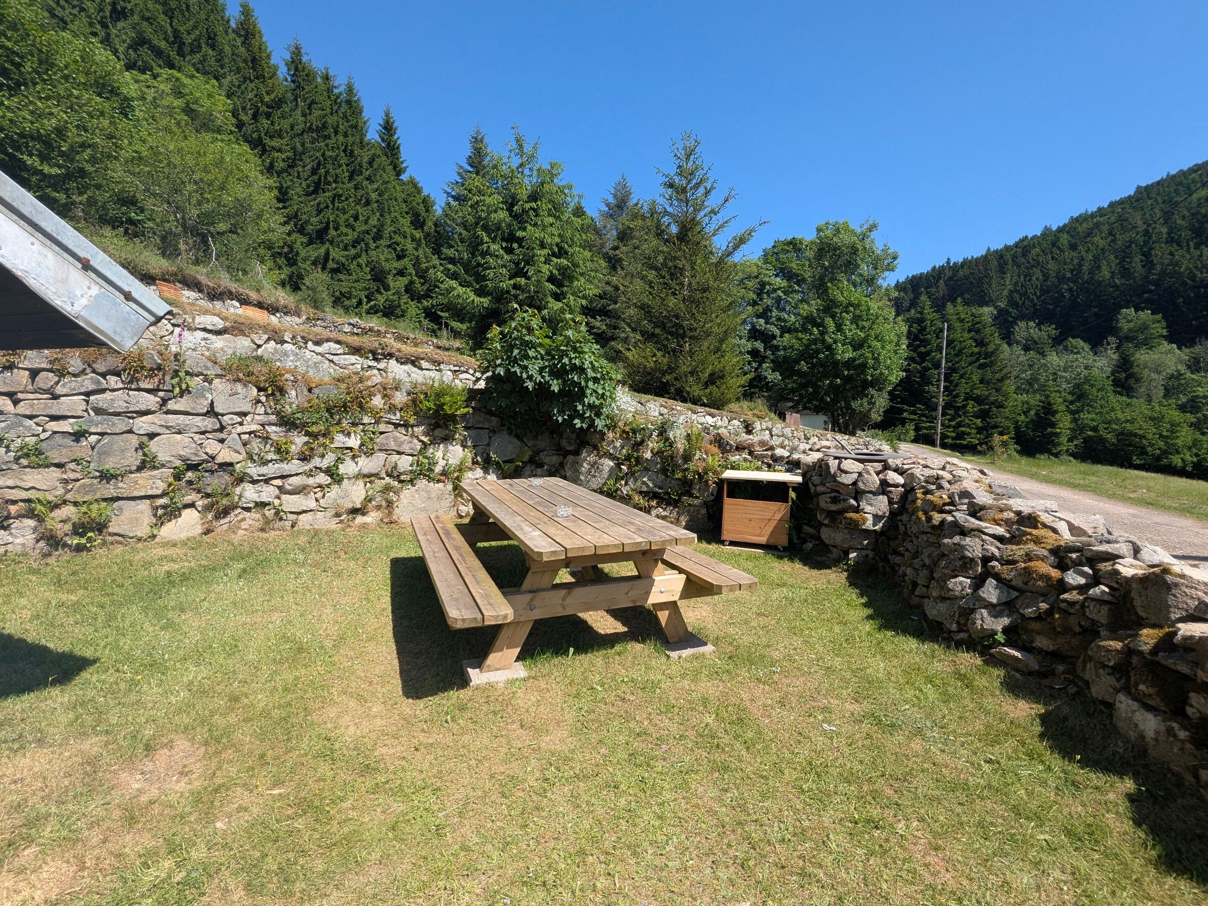 A picnic table with benches, a stone wall, and a grassy area with trees in the background.