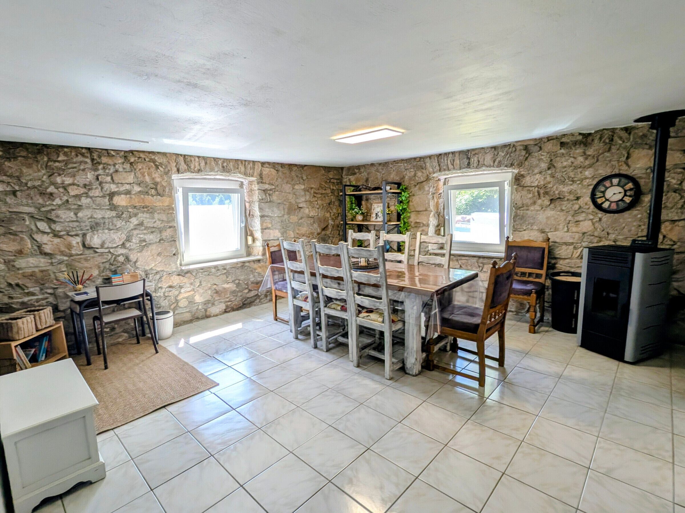 A stone-walled room with a tiled floor, a wooden dining table with chairs, a small white table, and a black heater.