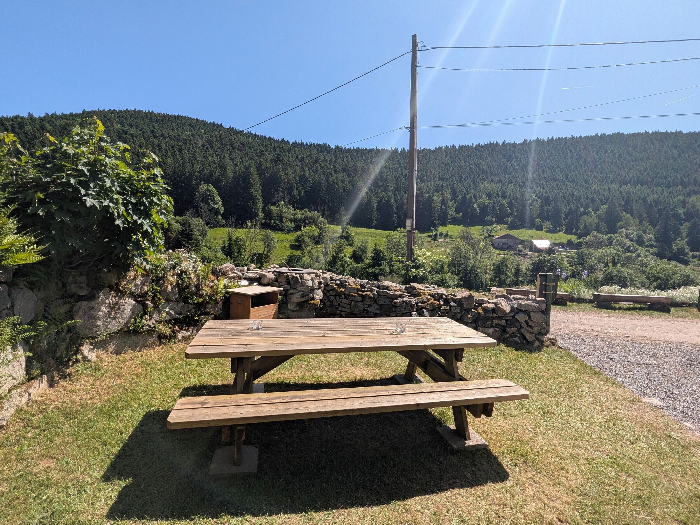 Uma mesa de piquenique com um muro de pedra ao fundo, situada numa área gramada com vista para uma colina arborizada.