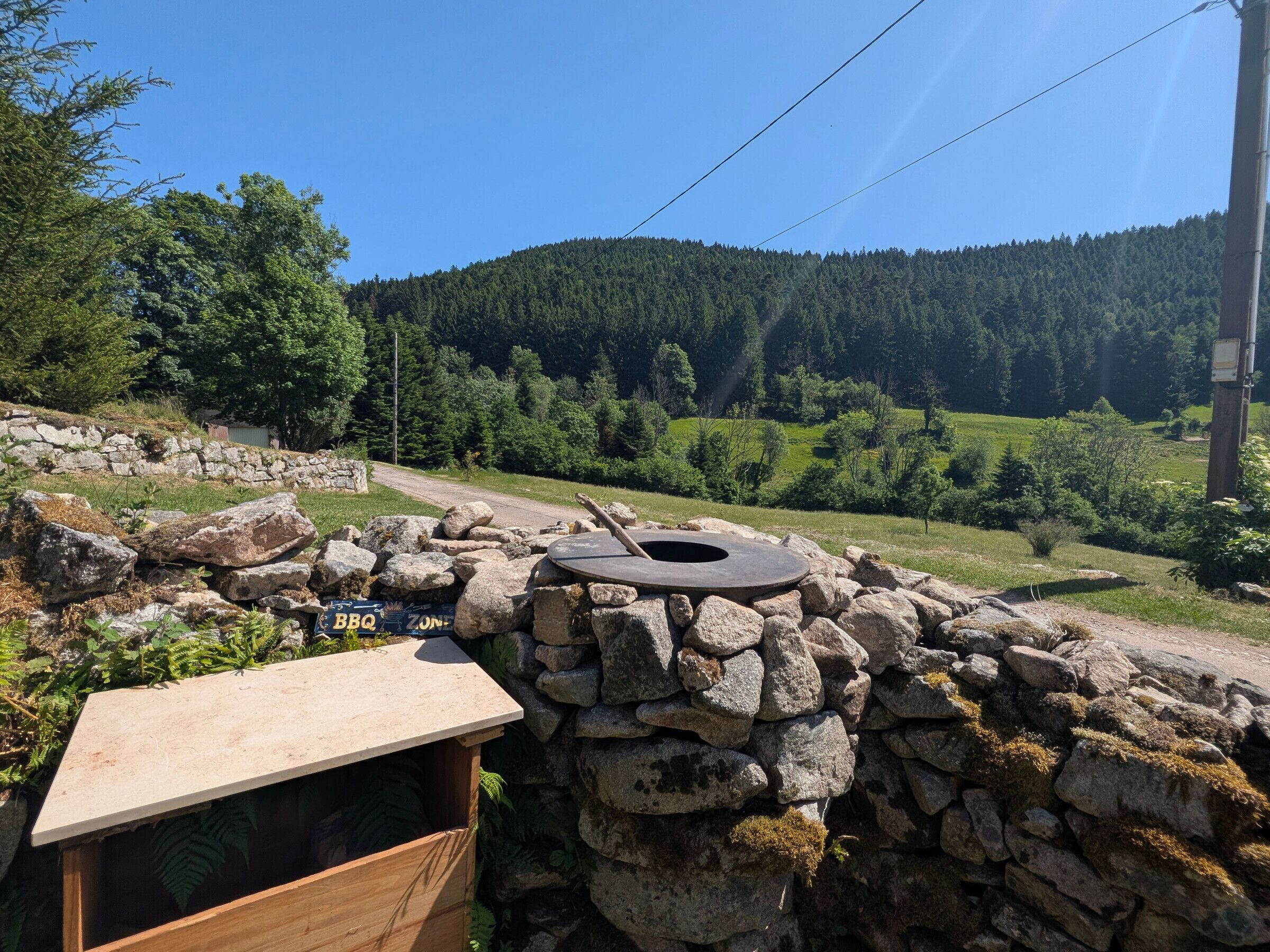 A stone wall with a BBQ grill and a wooden structure, set against a backdrop of a forested hillside.