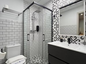 Un baño moderno con ducha envuelta en vidrio, paredes cubiertas de azulejos blancos, una pared de acento con diseño blanco y negro, un lavamanos blanco con grifo negro y un inodoro con tapa blanca.