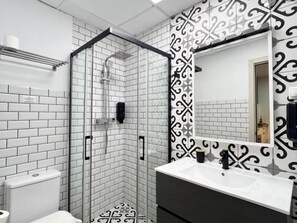 Ett badrum med en dusch som är avskärmad med glasväggar, vita kakelplattor i tunnelbanestil, en svartvit mönstrad vägg och en vit tvättställsskål med en mörk bänkskiva.