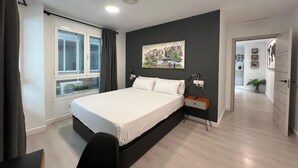 Ein modernes Schlafzimmer mit einem Bett, einem Fenster mit Vorhängen, einem Bild an der Wand und einem Flur, der in einen anderen Raum führt.