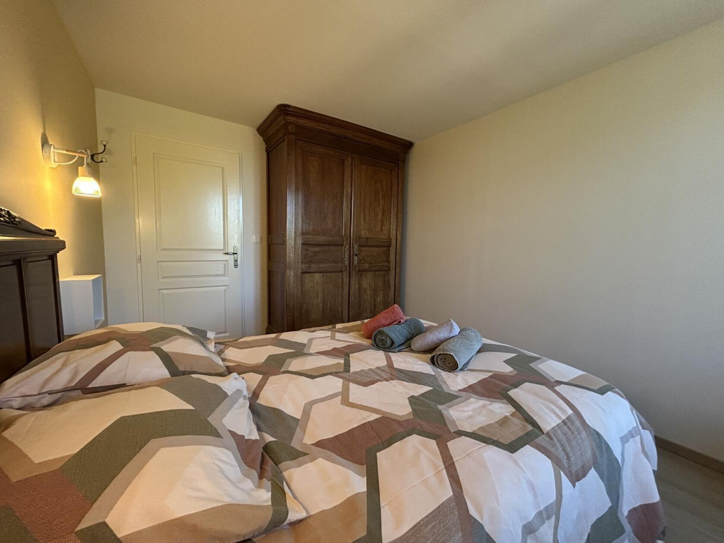 A bedroom with a bed, a wooden wardrobe, a door, and a wall-mounted light fixture.
