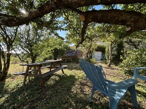 Un área de picnic con mesa y bancos de madera, rodeada de árboles y un pequeño edificio con techo de tejas.