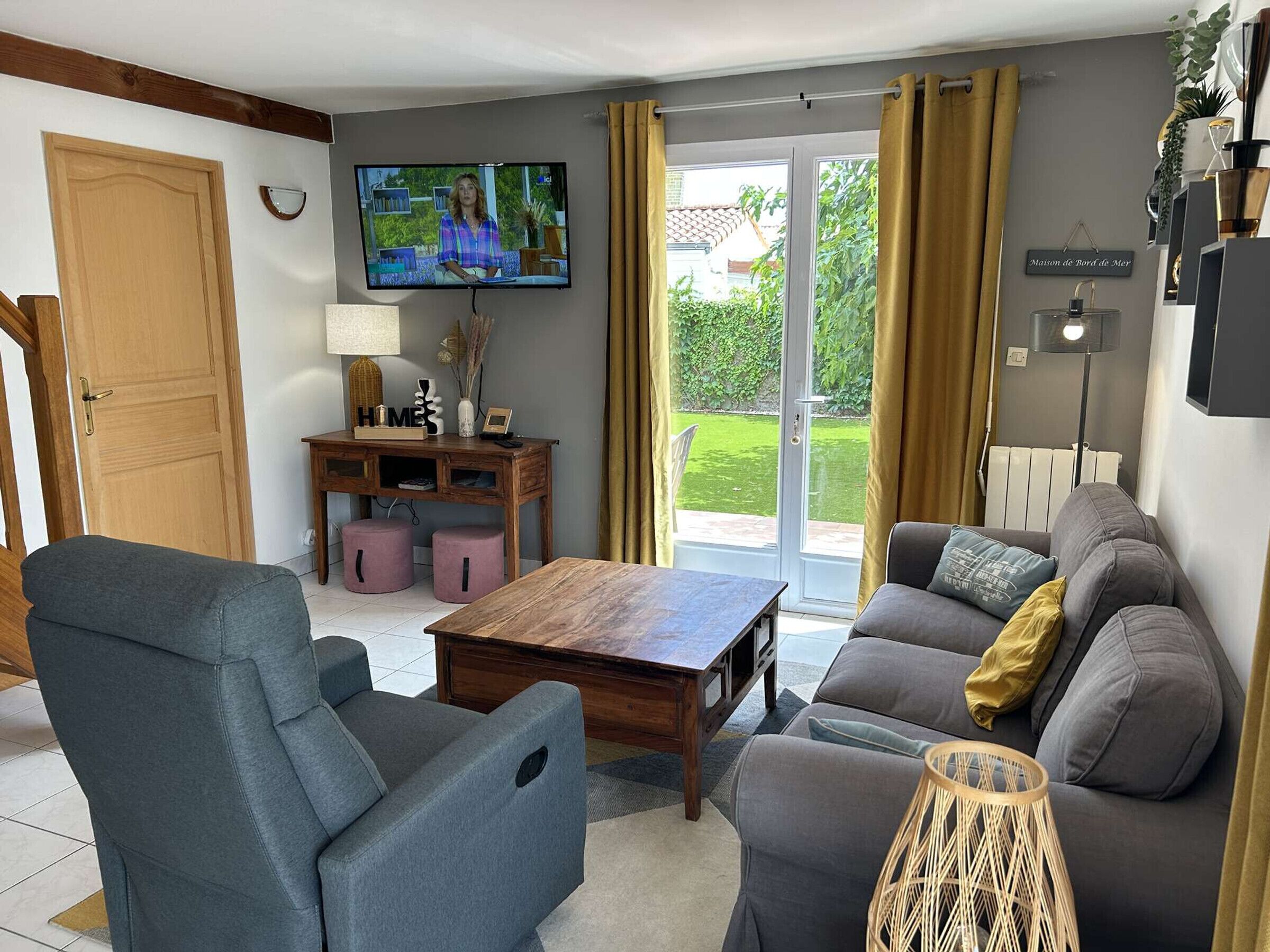 Un salon comprenant un téléviseur, un canapé, un fauteuil, une table basse et donnant sur un espace extérieur grâce à une porte vitrée.