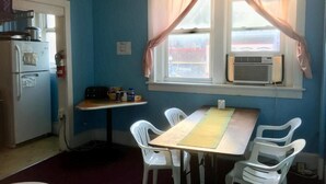 Een kleine keuken met een koelkast, fornuis en raam met gordijnen. Er staat een tafel met stoelen en een klein plankje met spullen.