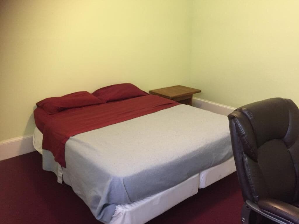 A single bed with a red and white bedspread, a wooden nightstand, and a black chair in a room with light green walls.