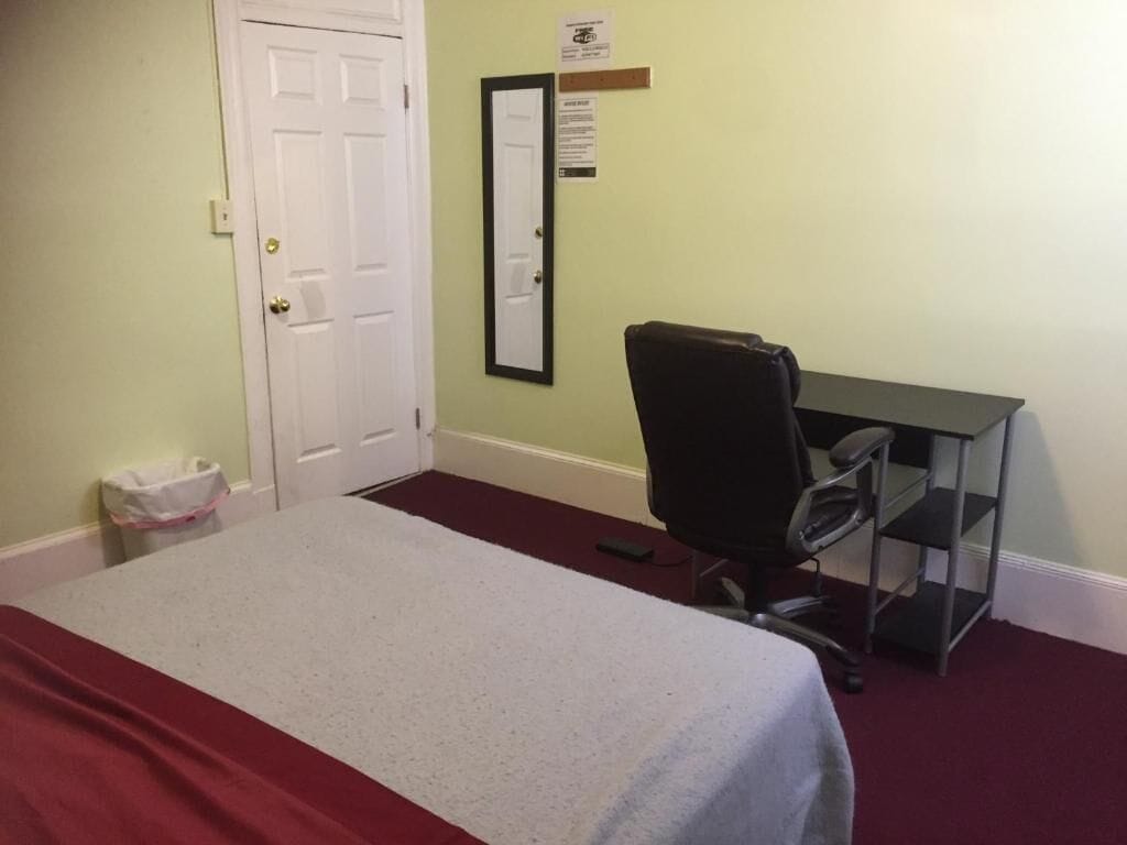 A room with a bed, a desk, a chair, and a trash bin.
