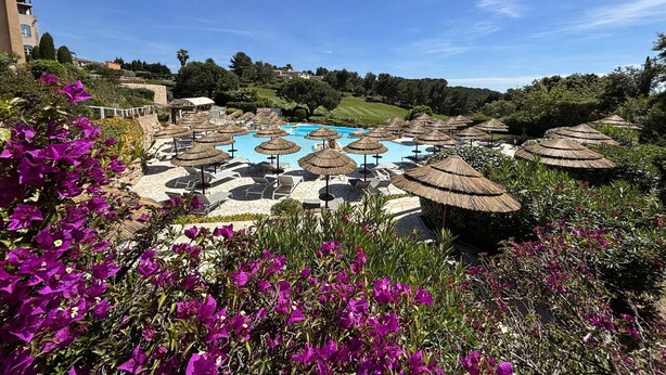 Zona de piscina amb gandules i ombrel·les, envoltada de vegetació i edificis.