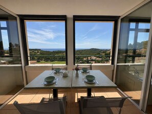 A dining table with four chairs, two windows with a view of a green landscape and buildings, and a balcony with a glass railing. - Apartment with AC, sea view - 6 guests (Saint-Cyr-sur-Mer)