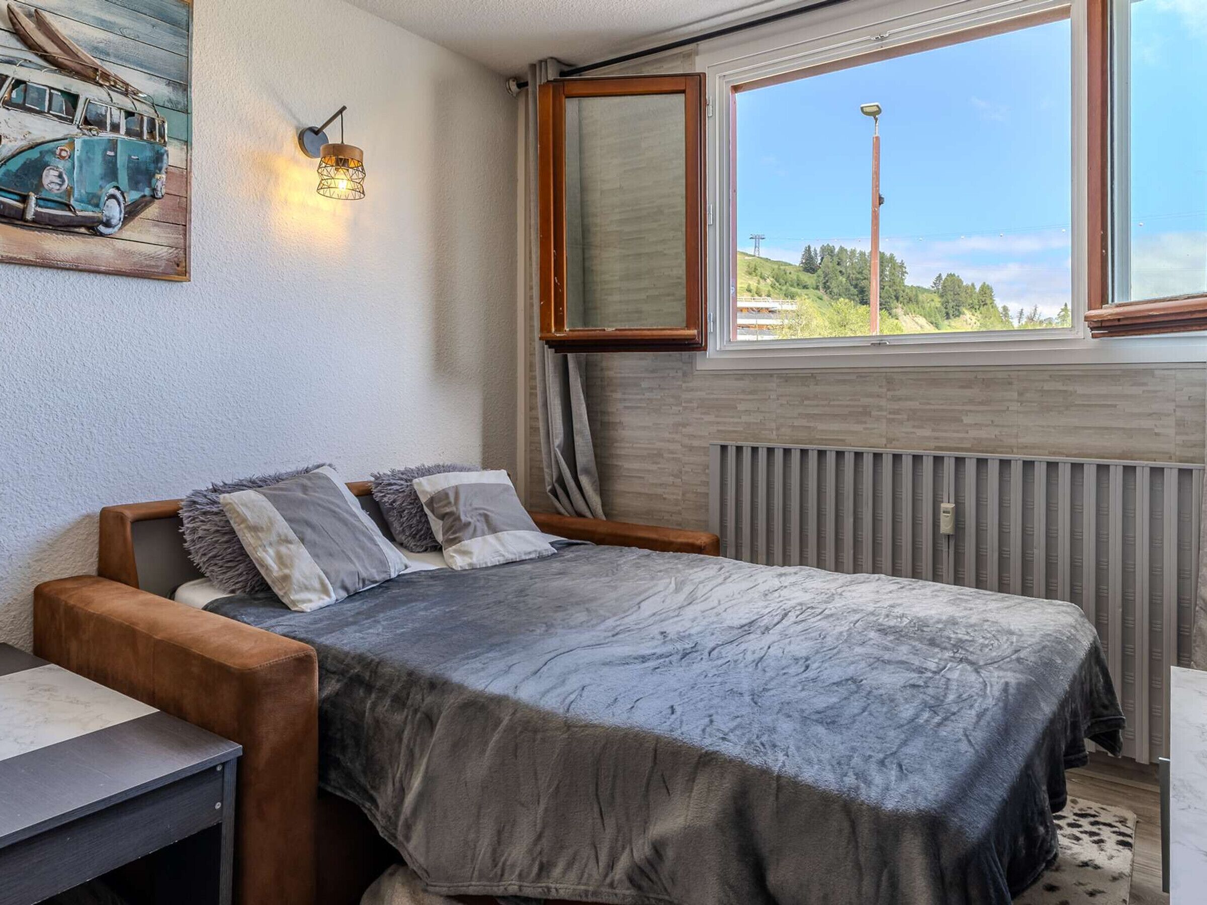 A bedroom with a bed, a wooden bench, a radiator, a window with a view, and a wall-mounted light fixture.
