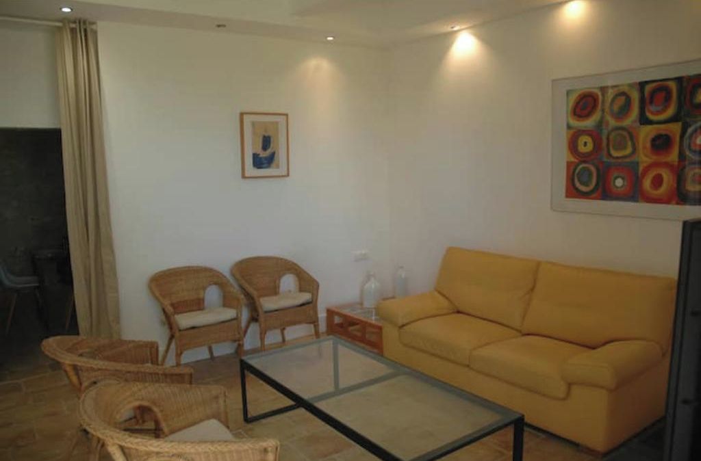 A living room with a yellow sofa, two wicker chairs, a glass coffee table, and a painting on the wall.