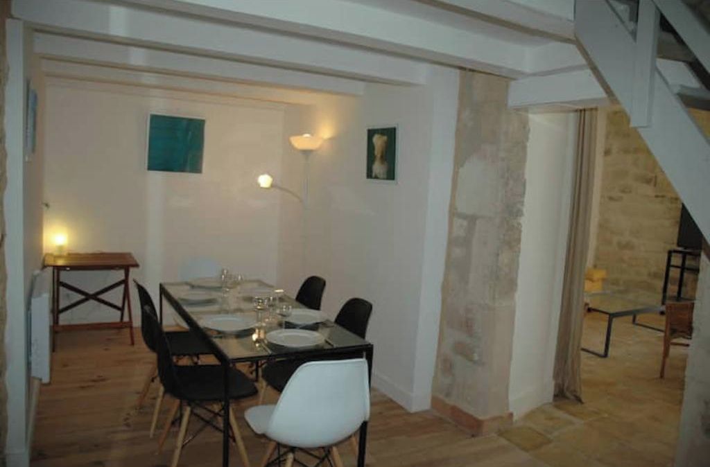 Un coin repas avec une table dressée pour quatre personnes, des chaises, une petite table avec une lampe et un escalier menant à un autre niveau.