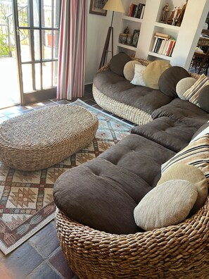 Ein gemütliches Wohnzimmer mit einer großen Eckcouch, zwei gewebten Hocker, einem gemusterten Teppich und einem Bücherregal mit Dekoartikeln.