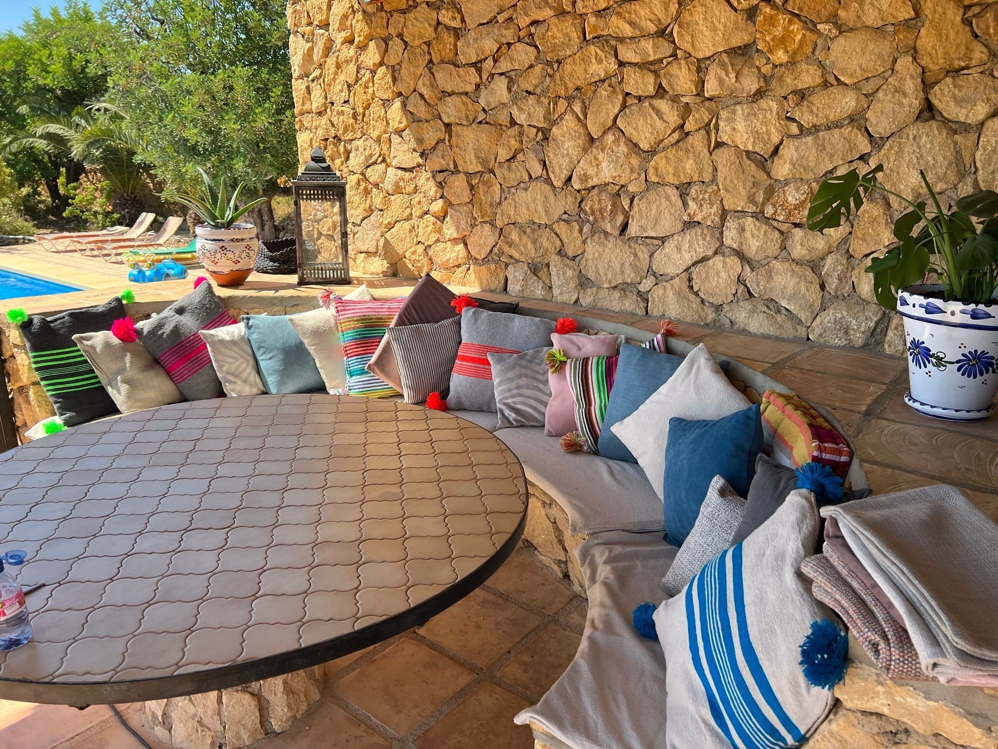 A stone wall, a round table, and a cushioned seating area with pillows and a potted plant.