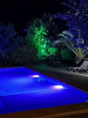 Ein Swimmingpool mit blauer Beleuchtung, umgeben von Bäumen und einer Liege.