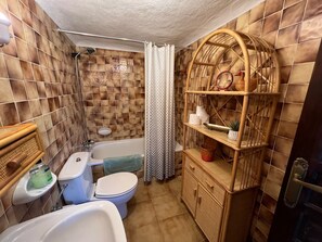 Un bany amb banyera, vàter i rentapans. Hi ha una cortina de bany i una prestatgeria amb articles de tocador.