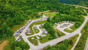 Vista aérea de um camping com veículos recreativos estacionados em áreas organizadas, cercado por uma floresta densa e uma rodovia.